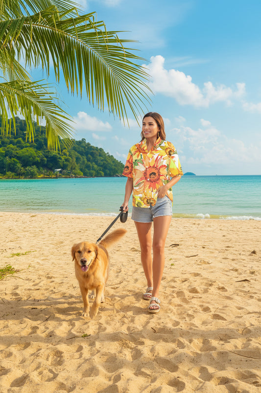 A woman wearing a short-sleeve button-up shirt with a large sunflower print, standing on a sandy beach with turquoise water and palm leaves in the background.