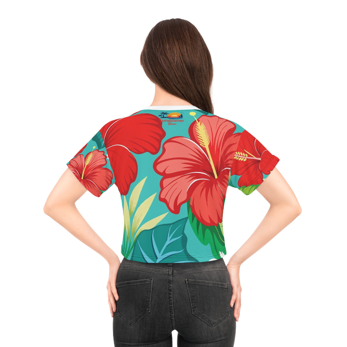 A woman shown from the back wearing a short-sleeve crop top with a bold tropical floral print featuring large red hibiscus flowers and green leaves on a teal background, paired with dark gray jeans against a plain white studio background.